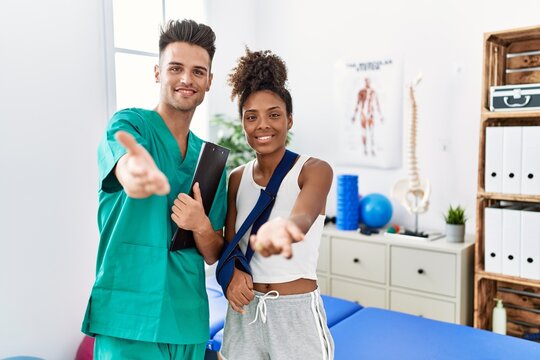 Physiotherapist working with patient wearing arm on sling at rehabilitation clinic smiling cheerful offering palm hand giving assistance and acceptance.