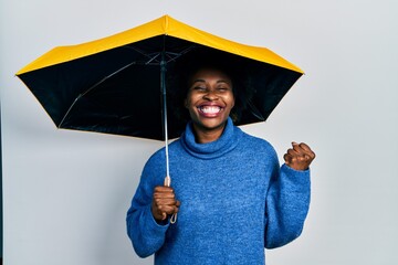 Young african american woman holding yellow umbrella screaming proud, celebrating victory and...