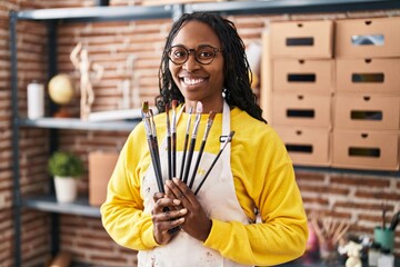 African american woman artist smiling confident holding paintbrushes at art studio