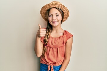 Beautiful brunette little girl wearing summer hat smiling happy and positive, thumb up doing excellent and approval sign