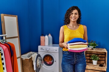 Young latin woman smiling confident holding folded clothes at laundry room