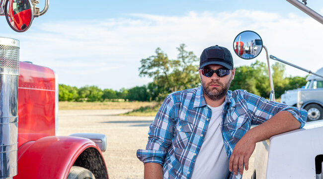 Handsome, professional looking truck driver wearing blue plaid shirt, protective sunglasses and black American baseball cap sits next to semi truck. Trucker is next to big rigs, looking at camera.