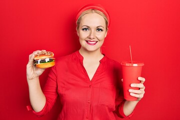 Young blonde woman eating a tasty classic burger with fries and soda smiling with a happy and cool smile on face. showing teeth.