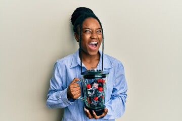 African american woman with braided hair holding food processor mixer machine with fruits angry and mad screaming frustrated and furious, shouting with anger looking up.