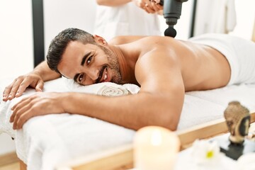 Obraz na płótnie Canvas Young hispanic man having back massage using percussion pistol at beauty center