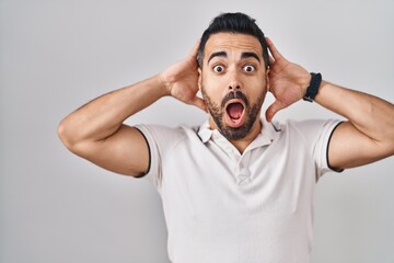 Young hispanic man with beard wearing casual clothes over white background crazy and scared with hands on head, afraid and surprised of shock with open mouth
