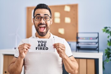 Young hispanic man with beard holding you are fired banner at the office celebrating crazy and amazed for success with open eyes screaming excited.