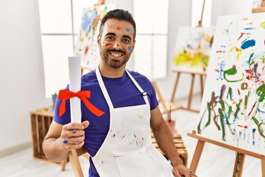 Young hispanic man smiling confident holding diploma at art studio
