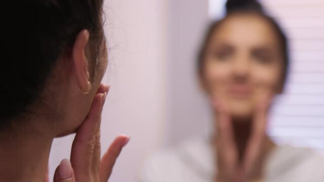 Rack focus from woman's reflection while rubbing face during skin care routine