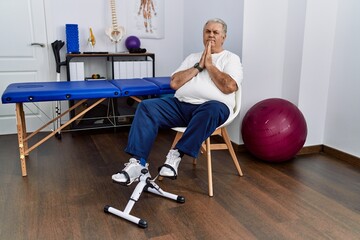 Senior caucasian man at physiotherapy clinic using pedal exerciser praying with hands together asking for forgiveness smiling confident.
