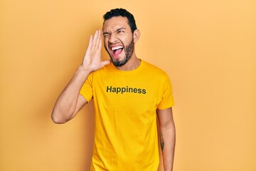 Hispanic man with beard wearing t shirt with happiness word message shouting and screaming loud to side with hand on mouth. communication concept.