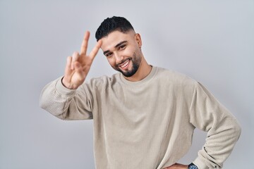 Fototapeta premium Young handsome man standing over isolated background smiling looking to the camera showing fingers doing victory sign. number two.
