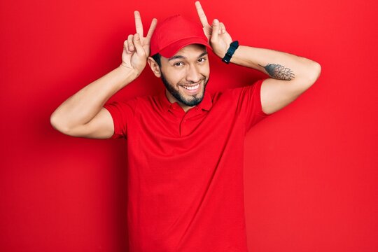 Hispanic man with beard wearing delivery uniform and cap posing funny and crazy with fingers on head as bunny ears, smiling cheerful