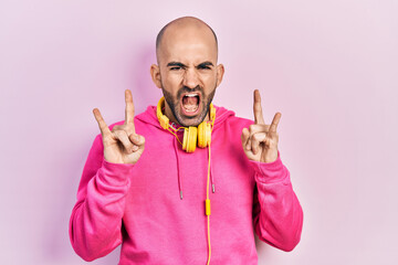 Naklejka premium Young bald man wearing gym clothes and using headphones shouting with crazy expression doing rock symbol with hands up. music star. heavy concept.