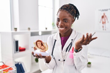 African american woman wearing doctor uniform holding anatomical model of uterus with fetus at...