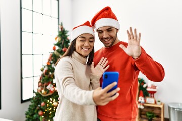 Young hispanic couple wearing christmas hat having video call by the smartphone at home.