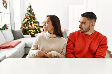 Young hispanic couple sitting on the table by christmas tree looking to side, relax profile pose with natural face with confident smile.