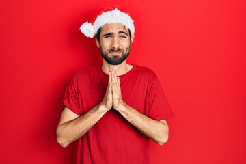 Young hispanic man wearing christmas hat praying with hands together asking for forgiveness smiling confident.