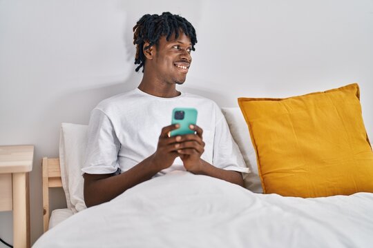African american man using smartphone sitting on bed at bedroom - Powered by Adobe