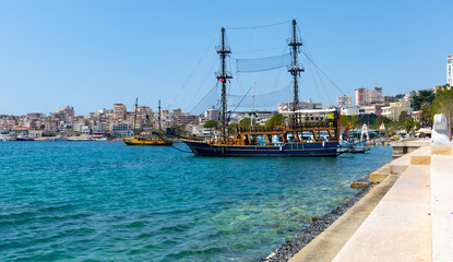 Scenic cityscape of Saranda on coast of gulf of Ionian Sea with vintage masted wooden sailing ships...