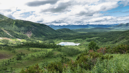 The lake in the valley is surrounded by mountains. There is lush green vegetation on the slopes. The blue sky with clouds is reflected in the water. Kamchatka. Vachkazhets. Lake Tahkoloch