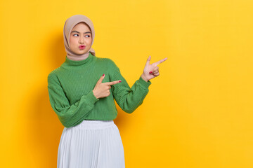 Beautiful Asian woman in green sweater pointing fingers aside at copy space isolated over yellow background