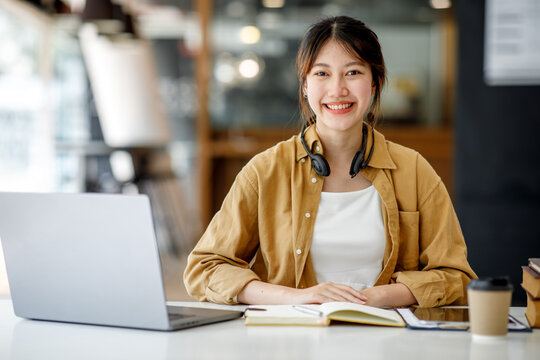 Young Adult Happy Smiling Hispanic Asian Student Wearing Headphones Talking On Online Chat Meeting Using Laptop In University Campus Or At Virtual Office. College Female Student Learning Remotely.