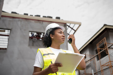 African female engineer using radio ordering work at construction project site with confidence.beautiful female worker with black skin,smart, leadership.PPE,helmet,safety,vest under construction house