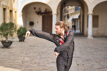 Fototapeta na wymiar Young man with beard and ponytail, wearing black transparent shirt with black polka dots and red roses, black pants and jacket, dancing flamenco in the city. Concept art, dance, culture, tradition.