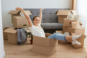 Portrait of positive happy delighted woman wearing white t shirt and jeans sitting in a cardboard box with raised arms and looking at camera, rejoicing her relocating to a new flat.