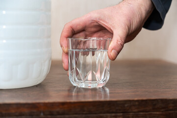 A man holds a glass of drinking water in his hand. A large white plastic bottle of fresh water is next to him on the table. Inside the room.