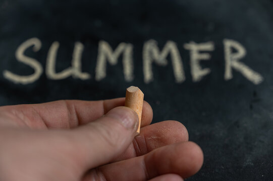 Close-up of a man's hand holding yellow chalk. The word SUMMER written on a black surface in the background. Handwritten text.