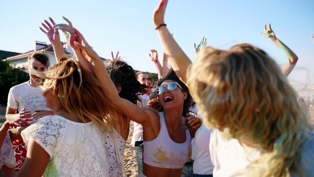 Cheerful people celebrate Holi festival stained in colored powder. Friends have fun at hindu holiday of spring, colors, love. Guys dance, jump on beach in slow motion. End of covid pandemic isolation.