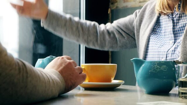 A conversation between a man and a woman over a cup of coffee. The woman is gesturing. Yellow cup and blue coffee pot. No faces, only hands