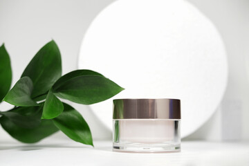 A mock up white jar of cream on a white gray background, on a white table with green leaves of a tropical plant, with hard shadows. Stylish look of the product, identity.