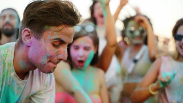 Cheerful people celebrate Holi festival stained in colored powder. Friends have fun at hindu holiday of spring, colors, love. Guys dance, jump on beach in slow motion. End of covid pandemic isolation.