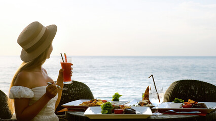 Young woman have romantic breakfast in sunrise at resort fine dining restaurant outdoor enjoy vacation looking at sea. Beautiful girl wear hat hold fruit cocktail. Healthy food drink for breakfast.