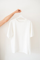 A woman holds a white T-shirt with a hanger against a light background.