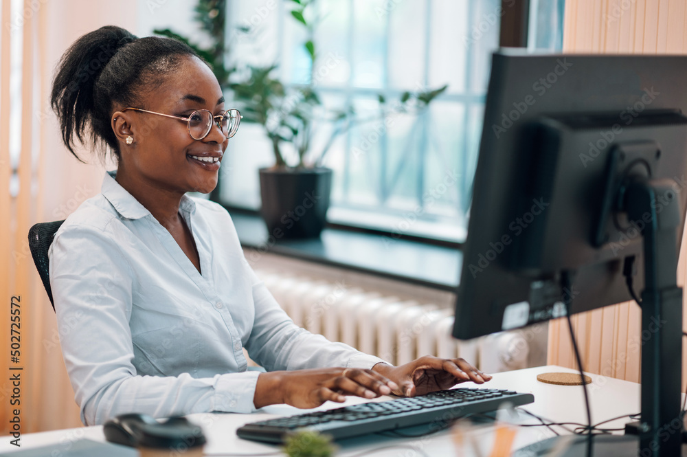 Wall mural Smiling millennial young black woman working on a computer in a office and consulting a client or a customer online. Confident businesswoman working on computer at her workplace at modern office.