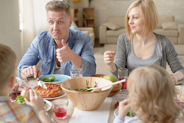 Beautiful Caucasian couple with kids sitting at dining table at home and eating appetizing lunch, father looking at son and showing thumb up