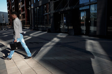 Obraz premium End of working day. Side view of African American manager with case moving away from modern business center, shadows seen on pavement