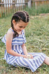 Fototapeta na wymiar A cute dark-haired girl with pigtails in a blue and white sundress holds a chicken in her hands while sitting on the lawn on a summer day.Childhood, summer, rural simple life, positive vibes.