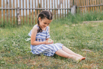 A cute dark-haired girl with pigtails in a blue and white sundress holds a chicken in her hands while sitting on the lawn on a summer day.Childhood, summer, rural simple life, positive vibes.