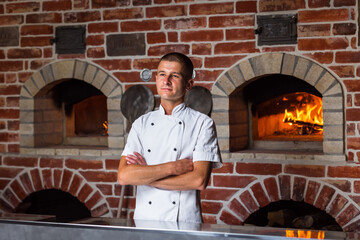 Portrait of a smiling pizza chef standing on the background of a wood-burning oven in the kitchen