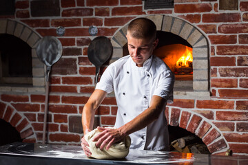 Chief chef kneads the dough on table with flour for cooking Italian pizza near a wood-burning oven...