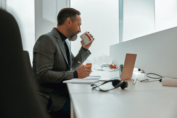 Side view of busy man sitting in modern office while drinking coffee and making notes on documents