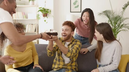 United multiracial group of young adult friends celebrating birthday party at home. Hipster brazilian man blowing out candles on cake
