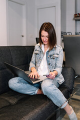 Woman using a laptop computer and a credit card while doing shopping online sitting on the sofa at home. Online shopping, online banking and e-commerce concept.