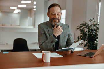 Smiling adult man in a jacket holding notepad while sitting in his office