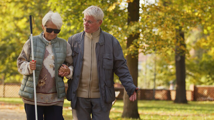 Life with disability. Elderly couple taking a walk in their favourite park in autumn. Disabled blind lady with white cane and sunglasses supported by her beloved husband. High quality photo